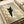 May include: Framed art print showing a black cat with bright yellow eyes peeking around what appears to be a corner or doorway, rendered in muted beige and olive green tones with soft shadowing. The wooden-framed piece sits on a light wooden surface next to rolled paper or fabric in neutral cream colors, creating a warm, minimalist styling scene.