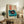 May include: Framed vintage-style poster featuring a turquoise record player with black vinyl and red label, displayed on rustic wooden sideboard alongside potted fern, ceramic vase, and stacked books in cozy living room with beige armchair and neutral walls