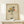 May include: Framed art print showing colorful wildflowers held in two hands, drawn in simple line art style with vibrant petals in orange, yellow, teal, and burgundy against cream textured background. Natural wood frame sits on light wooden surface beside small white wrapped gift with evergreen sprig. Soft natural lighting on neutral wall.