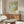 May include: Framed William Morris botanical print with sage green acanthus leaves and golden yellow flourishes displayed above a mid-century walnut credenza. Modern living room setting features cream upholstered chair, glass-top coffee table, and minimalist decor. Soft natural lighting creates warm shadows across neutral walls.