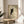 May include: Framed poster print featuring a black cat with golden yellow eyes peeking into view against a pale green background, displayed on a light wood round side table next to a beige upholstered chair with white pillow. Natural sunlight creates geometric shadows across the scene. The artwork shows the cat's head and partial neck with detailed whiskers and alert expression in a minimalist illustration style within a thin wooden frame.