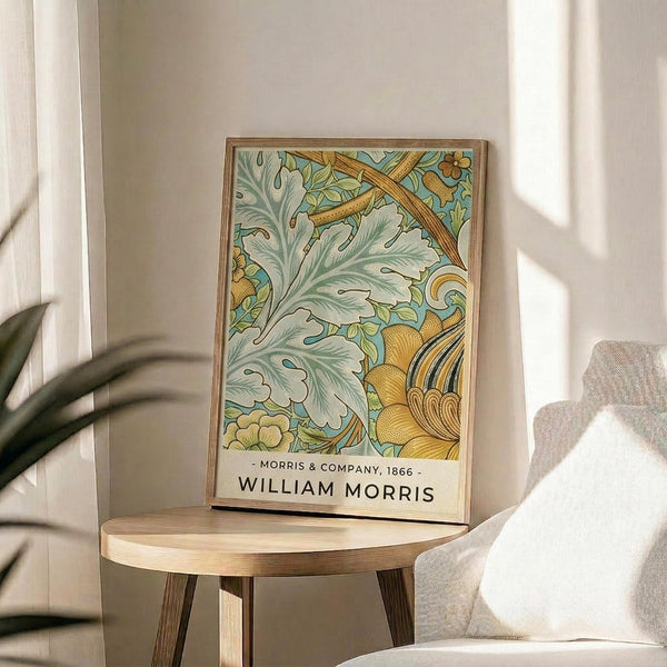 May include: Framed botanical art print featuring William Morris 1866 design with sage green acanthus leaves, golden yellow flowers and decorative elements on light blue background. Print displayed on round wooden side table next to beige upholstered chair with plant leaves visible in soft natural lighting against white walls.