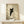 May include: Framed poster print featuring a black cat with bright yellow eyes peeking around what appears to be a corner or doorway, set against a soft beige background with geometric shadow patterns. The artwork is displayed in a thin wooden frame on a light wood shelf next to a small white wrapped gift with natural twine and dried botanical accent. Warm natural lighting creates diagonal shadows across the minimalist wall display.