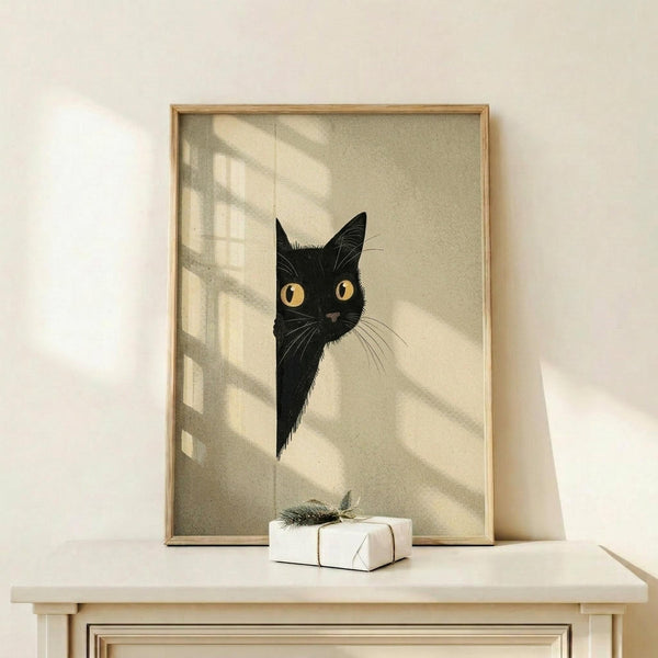 May include: Framed poster print featuring a black cat with bright yellow eyes peeking around what appears to be a corner or doorway, set against a soft beige background with geometric shadow patterns. The artwork is displayed in a thin wooden frame on a light wood shelf next to a small white wrapped gift with natural twine and dried botanical accent. Warm natural lighting creates diagonal shadows across the minimalist wall display.