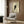 May include: Framed poster of black cat with bright yellow eyes peeking from behind white surface, displayed on wood-paneled wall above beige upholstered armchair with geometric pillow. Modern living room setting with brass floor lamp, side table with open book, and natural window light casting shadows across the minimalist artwork.