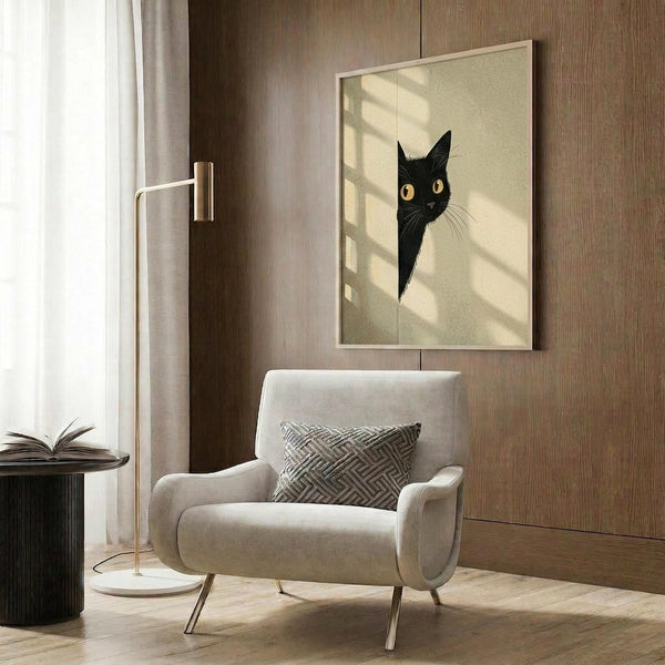 May include: Framed poster of black cat with bright yellow eyes peeking from behind white surface, displayed on wood-paneled wall above beige upholstered armchair with geometric pillow. Modern living room setting with brass floor lamp, side table with open book, and natural window light casting shadows across the minimalist artwork.