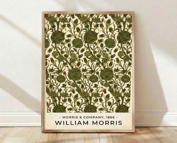 May include: Framed vintage botanical print with intricate green floral pattern on cream background, featuring detailed flowers and foliage in William Morris style. Light wood frame leaning against white wall on natural wood floor with soft natural lighting and shadows.