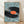 May include: Framed art print leaning against white wall on light wood floor. The poster shows an aerial view of a giant black vinyl record with orange label surrounded by tiny people casting long shadows on pale blue ground. A realistic turntable arm sits on the record's edge. The artwork has a surreal, miniature photography style in a thin light wood frame.