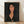 May include: Framed portrait artwork in the style of Amedeo Modigliani featuring a woman with elongated face, dark hair, and melancholic expression against black background. She wears dark clothing with a small rose detail. The print has warm flesh tones and is displayed in a thin wooden frame leaning against white wall on light wooden flooring with natural shadows.