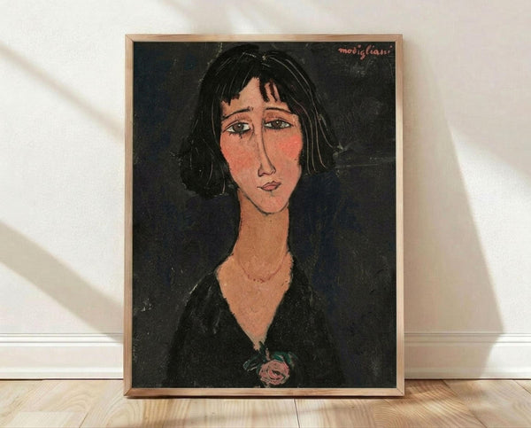 May include: Framed portrait artwork in the style of Amedeo Modigliani featuring a woman with elongated face, dark hair, and melancholic expression against black background. She wears dark clothing with a small rose detail. The print has warm flesh tones and is displayed in a thin wooden frame leaning against white wall on light wooden flooring with natural shadows.