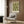 May include: Framed botanical poster with green floral pattern on cream background hanging on wood-paneled wall above beige upholstered armchair with geometric pillow. Modern floor lamp with brass finish and marble base stands nearby next to round black side table with open book. Warm neutral interior with curtained window.