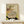 May include: Japanese woodblock print artwork in wooden frame showing striped cat drinking from decorative water basin with goldfish, pineapples visible in background, displayed on light wood shelf next to small white wrapped gift against cream wall
