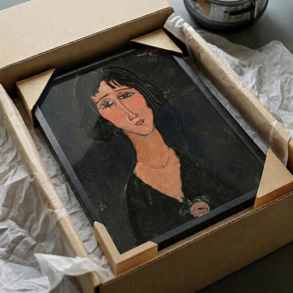 Modigliani-style portrait painting in a black frame, packed in cardboard shipping box with protective tissue paper. The artwork shows a woman with elongated neck, dark hair, and characteristic stylized features in muted tones against a dark background. Wooden frame corners and magnifying glass visible nearby on white surface.