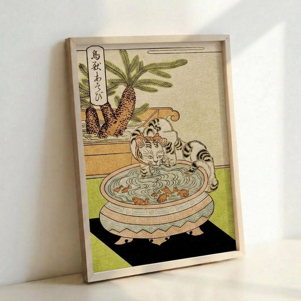 Japanese woodblock print style artwork showing a striped cat drinking from an ornate ceramic bowl filled with water and small fish. A potted palm tree sits in the background. The image features muted earth tones with green accents and includes Japanese calligraphy in the upper left corner. The artwork is displayed in a light wooden frame leaning against a white wall.