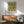 May include: Green and cream botanical pattern poster with "Morris & Company, 1866 William Morris" text, displayed in thin wooden frame above modern walnut sideboard. Mid-century modern living room setting with sage green upholstered armchair, white ceramic table lamp, and natural lighting creating wall shadows.