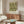 May include: Framed botanical print with green floral pattern on cream background hanging above wooden sideboard in minimalist living room. White curved chair and oval glass coffee table with colorful abstract art visible. Warm wood tones, neutral walls, and modern Scandinavian-style furniture create serene interior setting.