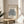 May include: Framed art print featuring blue botanical cutout shapes resembling coral or seaweed against cream background, displayed on round wooden side table next to beige upholstered chair. Natural sunlight creates geometric shadows on white wall. Green plant leaves visible in corner. Minimalist Scandinavian-style interior setting with neutral tones and natural wood frame.