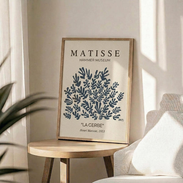 May include: Framed art print featuring blue botanical cutout shapes resembling coral or seaweed against cream background, displayed on round wooden side table next to beige upholstered chair. Natural sunlight creates geometric shadows on white wall. Green plant leaves visible in corner. Minimalist Scandinavian-style interior setting with neutral tones and natural wood frame.