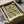 May include: Vintage botanical poster with intricate green floral pattern featuring detailed leaves, vines, and circular flower motifs on cream background. Black-framed print sits in open cardboard shipping box with white tissue paper and wooden tube packaging materials visible around it on neutral surface.