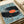 May include: Framed art print showing a large black vinyl record with orange center label and tonearm, surrounded by tiny people figures casting shadows on light blue background, displayed on white bedding next to rolled paper in soft natural lighting
