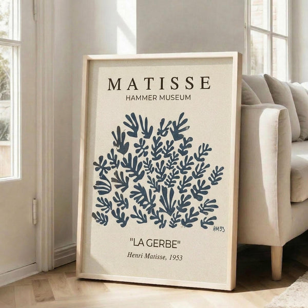 May include: Framed poster with cream background featuring dark blue botanical cut-out shapes resembling coral or seaweed, displayed leaning against white wall next to beige upholstered chair in bright room with large windows and wooden floors.
