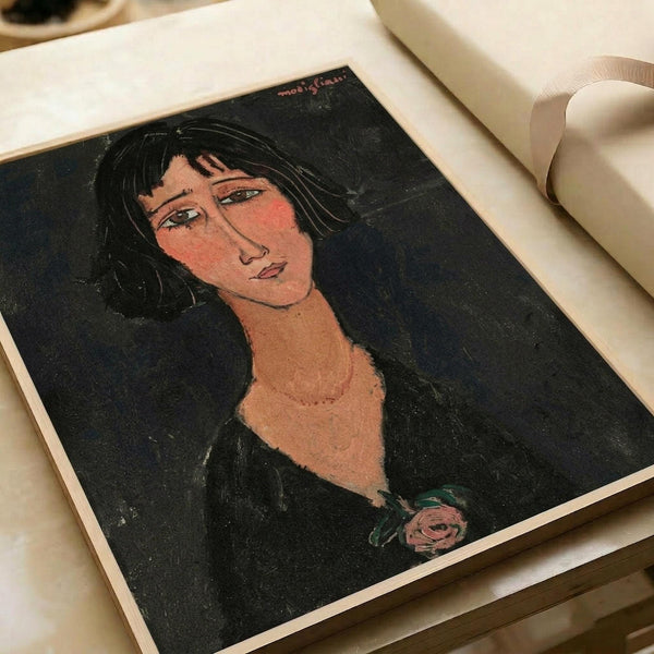 Portrait of a woman with dark hair and elongated neck in Modigliani's distinctive style, featuring warm peachy skin tones against a dark background with a small pink rose detail. The artwork is displayed as a print in an open art book or portfolio with cream-colored pages and white ribbon visible at the edges.