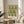 May include: Framed botanical print with green floral pattern on cream background displayed on round wooden side table next to beige upholstered chair with geometric patterned throw pillow. Natural sunlight streams across neutral-toned room with plant leaves visible in corner. Print features intricate repeating design of stylized flowers and foliage in olive green tones within thin wooden frame.