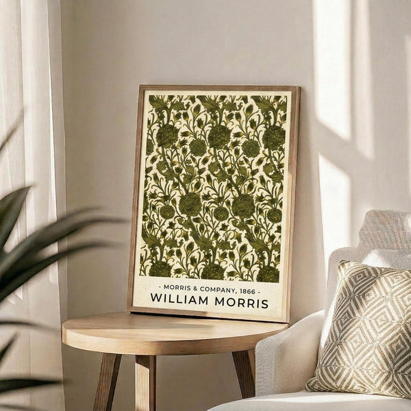 May include: Framed botanical print with green floral pattern on cream background displayed on round wooden side table next to beige upholstered chair with geometric patterned throw pillow. Natural sunlight streams across neutral-toned room with plant leaves visible in corner. Print features intricate repeating design of stylized flowers and foliage in olive green tones within thin wooden frame.