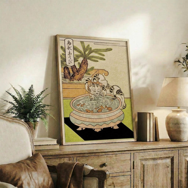 May include: Framed Japanese woodblock print featuring a striped cat drinking from an ornate basin, with pine trees and Japanese text in background. The artwork displays traditional ukiyo-e style with muted earth tones and green accents. Print is displayed on rustic wooden sideboard alongside fern plant, books, and ceramic vase in neutral-toned living room setting.