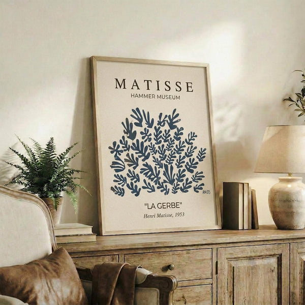 May include: Framed vintage-style art print featuring dark blue botanical cutout shapes resembling coral or seaweed on cream background, displayed on rustic wooden sideboard alongside fern plant, books, and ceramic vases in neutral-toned living room setting with upholstered furniture visible.