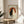 May include: Framed artwork displaying two cats - one black, one white - sitting together against an orange circular background with Japanese characters, styled in traditional ukiyo-e art with weathered textures. The wooden-framed print sits on a round wooden side table next to a beige upholstered chair, with soft natural lighting and plant shadows visible in a minimalist room setting.