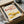 May include: Framed artwork showing two colorful lobster illustrations on cream background - one pink with multicolored spots, one yellow with red and blue dots. Black frame sits in cardboard shipping box surrounded by white tissue paper and wooden packing materials on white surface.