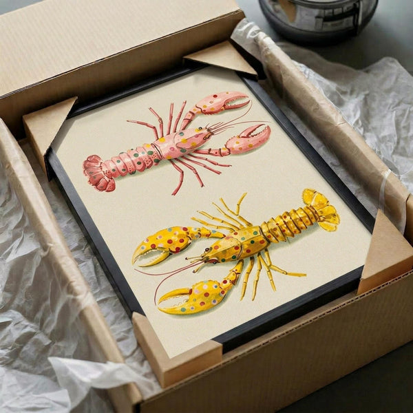 May include: Framed artwork showing two colorful lobster illustrations on cream background - one pink with multicolored spots, one yellow with red and blue dots. Black frame sits in cardboard shipping box surrounded by white tissue paper and wooden packing materials on white surface.