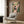 May include: Framed poster print mounted on wood-paneled wall above cream upholstered armchair with geometric pillow. Artwork features black cat reading newspaper surrounded by lush green plants on coral pink background. Modern reading nook with brass floor lamp, dark round side table with open book, and natural lighting from window with sheer curtains.