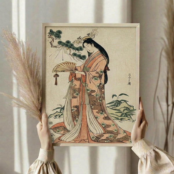 May include: Hands holding a framed Japanese woodblock print showing a woman in elaborate orange and cream kimono with floral patterns, holding a decorative fan. The artwork features traditional ukiyo-e style with pine tree branches above and plants below. Dried pampas grass visible on left side, neutral beige wall background.