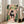 May include: Framed poster print leaning against wall next to beige sofa in bright living room with white walls and large windows. Artwork shows black cat with yellow eyes reading newspaper while sitting on pink toilet, surrounded by lush green houseplants including trailing pothos and snake plants. Pink bathroom setting with coral walls and botanical wall art. Light wood frame.
