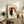 May include: Framed artwork featuring two cats - one black, one white - sitting back-to-back against a rust-colored circular background with Japanese text, displayed on a rustic wooden console alongside books, a ceramic vase, and a fern plant in a cozy living room setting with neutral tones and natural lighting.