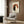 May include: Japanese-style artwork featuring two cats silhouettes - one black, one white - sitting back-to-back against an orange-red circular background with Asian characters, displayed in light wood frame on wood-paneled wall above beige upholstered chair with geometric patterned pillow, brass floor lamp and side table with open book nearby