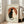 May include: Framed poster displaying two cats silhouetted against an orange-red circle resembling a sun or moon, with Japanese characters surrounding the design. The black cat and white cat sit back-to-back in a yin-yang composition on aged, textured paper background. Light wood frame leans against white wall next to beige upholstered chair in bright, modern living room with large windows.