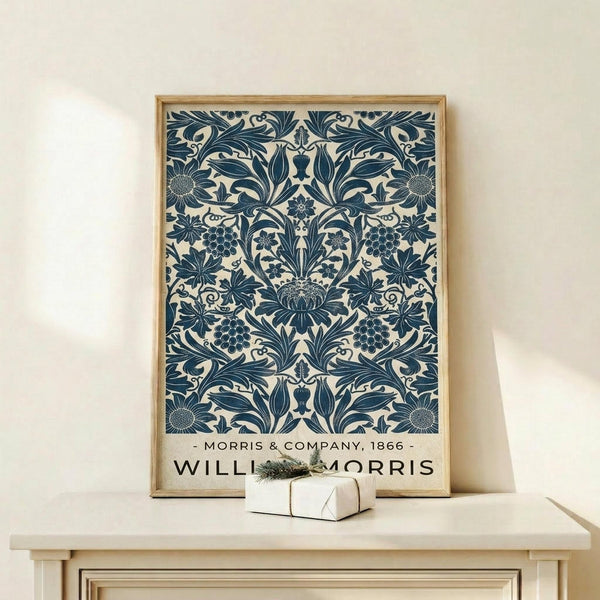May include: Framed vintage botanical poster with intricate blue floral pattern on cream background, featuring ornate Victorian-style damask design with leaves, flowers, and grape clusters. Natural wood frame displayed on light wooden console table beside small white wrapped gift with greenery sprig against neutral wall.