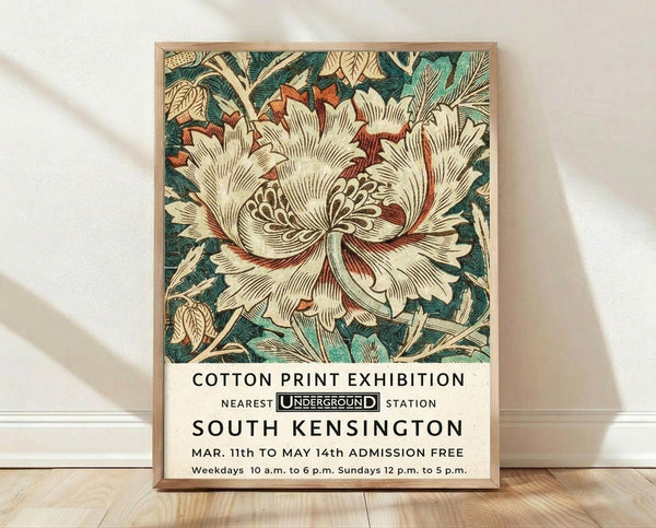 May include: Framed vintage botanical poster leaning against white wall on wooden floor. Features large cream peony flower with red accents and teal-green foliage in William Morris style. Black text below reads "Cotton Print Exhibition" with Underground logo and "South Kensington" location details. Natural wood frame, soft fabric shadows visible on textured white wall background.