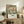 May include: Framed vintage-style botanical poster featuring large cream peony with red accents and teal foliage, displaying exhibition text for South Kensington. Light wood frame sits on rustic wooden sideboard beside fern plant, ceramic vase, and books. Beige armchair with brown leather cushion visible in foreground against white wall in cozy living room setting.