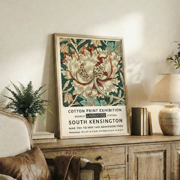 May include: Framed vintage-style botanical poster featuring large cream peony with red accents and teal foliage, displaying exhibition text for South Kensington. Light wood frame sits on rustic wooden sideboard beside fern plant, ceramic vase, and books. Beige armchair with brown leather cushion visible in foreground against white wall in cozy living room setting.