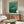 May include: Japanese-style artwork depicting an orange and black striped tiger swimming through turquoise green waves with white foam, displayed in a thin gold frame above a wooden sideboard. Modern living room setting with cream upholstered chair, brass floor lamp, white table lamp, oval glass coffee table, and white walls with natural lighting.