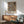 May include: Framed William Morris botanical poster with intricate floral pattern in deep navy, coral pink, and cream tones displayed above a mid-century modern walnut credenza. Green upholstered armchair with wooden arms sits nearby, along with a round wooden side table and white ceramic table lamp. Bright natural light creates geometric shadows on the white wall in this minimalist living room setting.