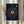May include: Navy blue framed poster with gold vinyl record design and spiral song lyrics text, displayed on white surface next to dried pampas grass stems against neutral curtained background