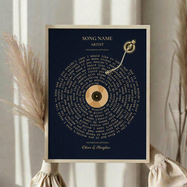 May include: Navy blue framed poster with gold vinyl record design and spiral song lyrics text, displayed on white surface next to dried pampas grass stems against neutral curtained background