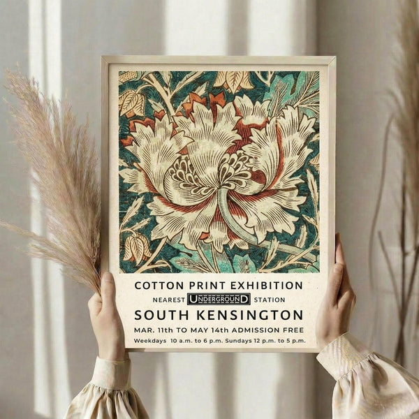May include: Hands holding a framed poster featuring a large decorative cream and rust-colored flower with intricate petals surrounded by teal and green foliage in vintage botanical style. Black text below reads "COTTON PRINT EXHIBITION" with Underground logo and "SOUTH KENSINGTON" venue details. Light wood frame against white curtained background with dried pampas grass visible on left side.