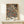 May include: Framed botanical poster with intricate floral pattern on navy background, featuring cream and coral flowers, ornate leaves, and decorative vines in William Morris style. Golden wood frame sits on light wooden shelf next to small white gift box with evergreen sprig. Cream colored wall backdrop creates minimalist display setting.