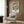 May include: Framed vintage botanical poster featuring ornate cream and teal floral design hung on wood-paneled wall above beige upholstered armchair with geometric pillow. Modern brass floor lamp and dark round side table with open book complete the neutral-toned reading nook scene.